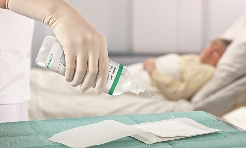 V popředí ruka s lékařskou rukavicí nalévá oplachový roztok Prontosan na podložku položenou na stole vedle tuby s Askina Calgitrol. V pozadí leží pacientka na nemocničním lůžku.