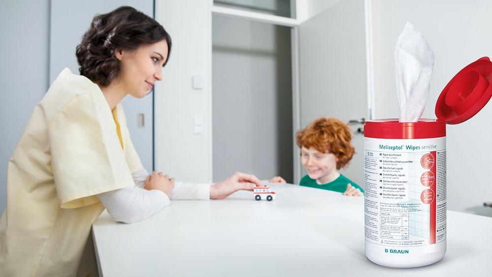 Sestra stojí u stolu před malým usmívajícím se chlapcem. Na stole je malý model sanitky, kterou tlačí žena, na tu se dívá chlapec. V popředí balení ubrousků Meliseptol Wipes.