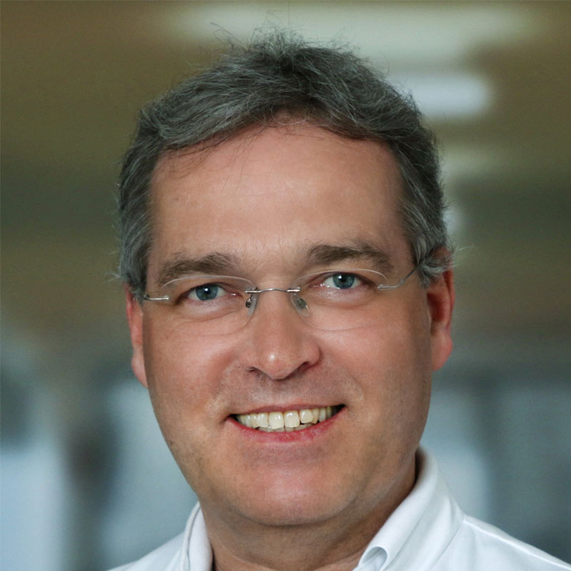 Portrét profesora doktora Markuse Gollinga v doktorském plášti, který se sebevědomě usmívá do kamery